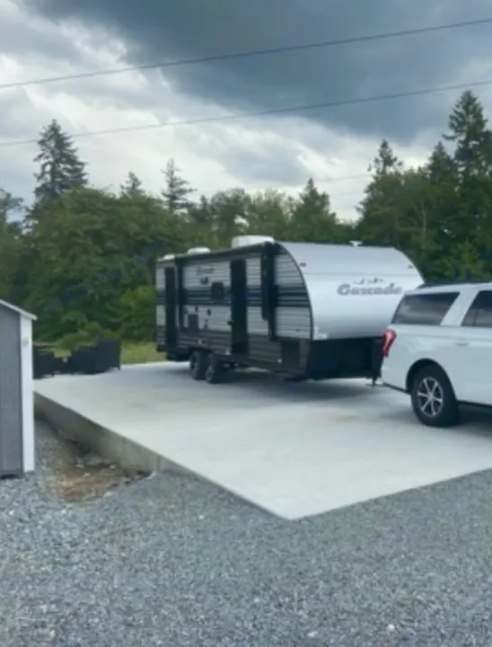 Modern RV rental travel trailer on a concrete pad, hitched to an SUV. Surrounded by trees under a cloudy sky.. Forest River Cherokee Grey Wolf 2021