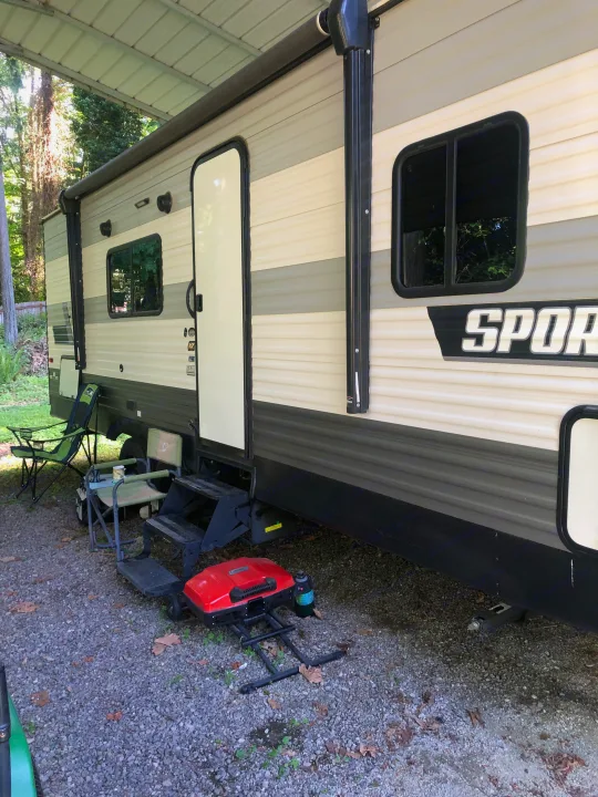 This RV rental at a gravel campsite features camping chairs, steps, and a portable grill, ready for outdoor fun.. KZ Sportsman SE 2022