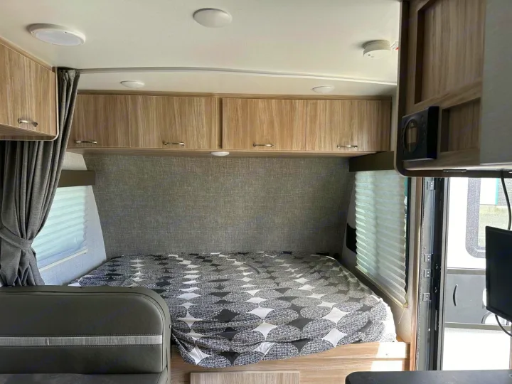 This RV rental bedroom features a patterned bed, light wood overhead cabinets, and a window for comfortable, compact travel.. Winnebago Micro Minnie 2022