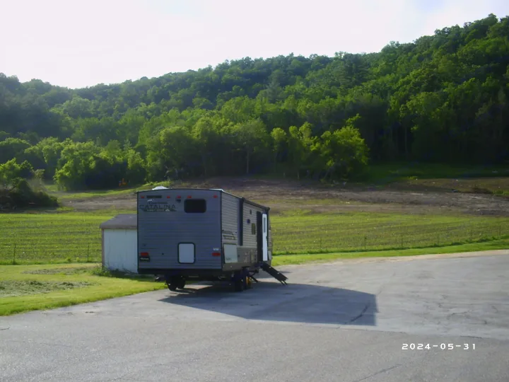 Grey RV rental travel trailer parked on asphalt, surrounded by green fields and a wooded hill.. COACHMAN CATALINA 261BHS 2024