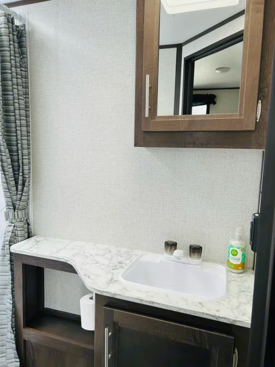 This clean RV rental bathroom features a functional vanity with sink, mirror, storage, and patterned curtain.. Dutchmen Coleman Light 2021