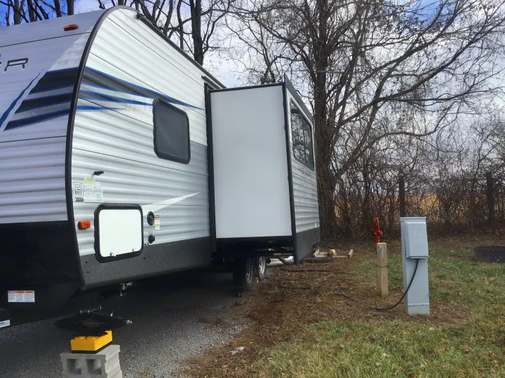 This RV rental shows a travel trailer with an extended slide-out at a gravel campsite, hooked up to utilities near bare trees.. Prime Time Avenger 2021