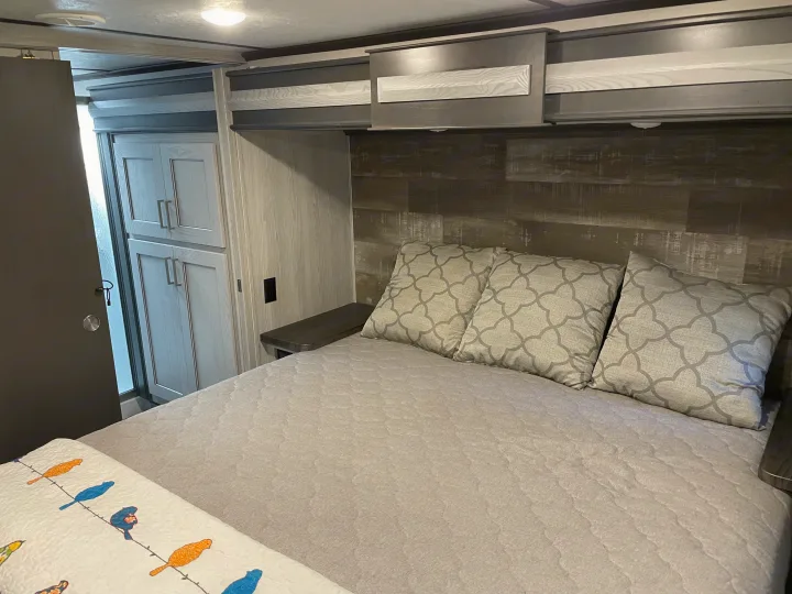 A cozy RV rental bedroom awaits, featuring a comfortable bed with patterned pillows, overhead storage, and ample light.. Keystone RV Montana High Country 2021
