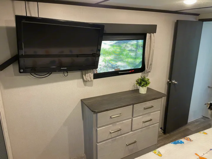 Cozy RV rental bedroom features a wall-mounted TV, window, light gray dresser, and plant.. Keystone RV Montana High Country 2021