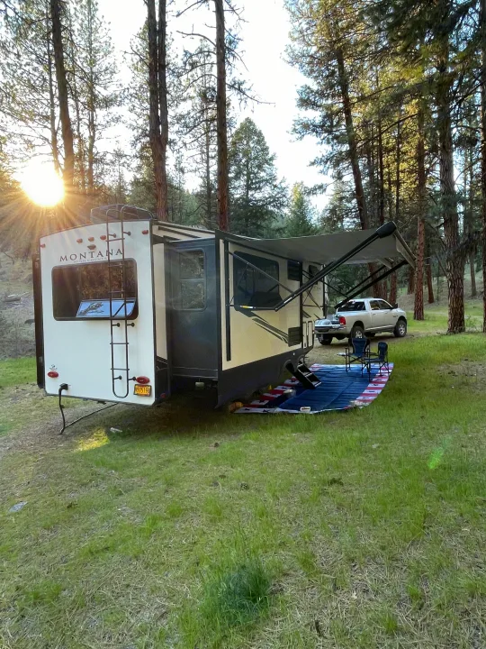 Serene forest scene: an RV rental with awning, parked next to a truck on grass. Camping chairs await outdoor enjoyment.. Keystone RV Montana High Country 2021