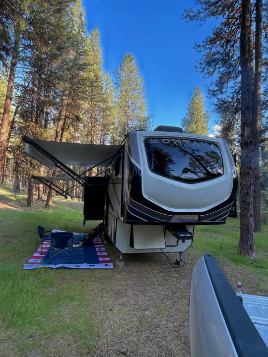 Enjoy nature with this large fifth-wheel RV rental, perfectly set up at a forest campsite with an awning & patriotic mat.. Keystone RV Montana High Country 2021