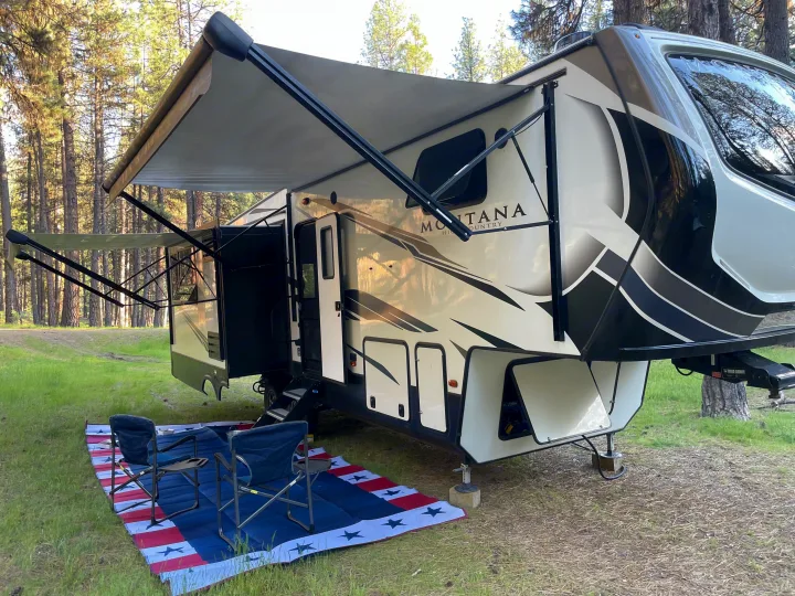 Spacious Montana fifth-wheel RV rental in a serene forest, with an extended awning, outdoor mat, and chairs, perfect for camping.. Keystone RV Montana High Country 2021
