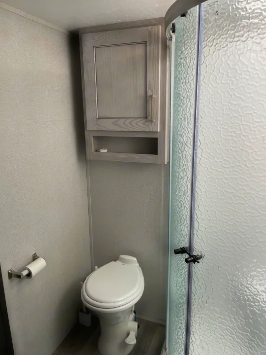Compact RV rental bathroom featuring a white toilet, overhead cabinet, and a textured glass shower enclosure.. Keystone RV Montana High Country 2021