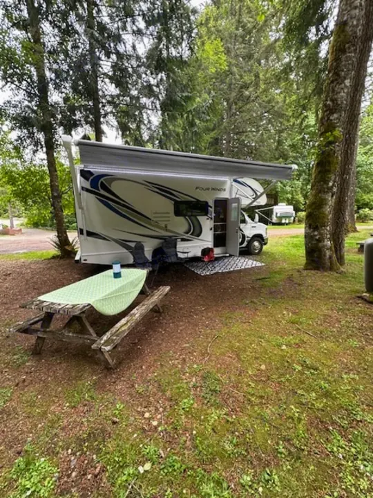 This RV rental is set up at a leafy campground, featuring a motorhome with an extended awning and a picnic table.. Thor Four Winds 2021