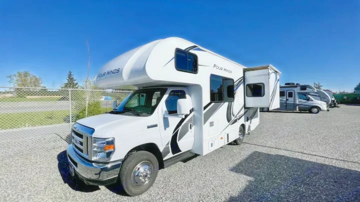 This white Four Winds Class C RV rental features an extended slide-out, parked on a gravel lot under blue sky.. Thor Four Winds 2021
