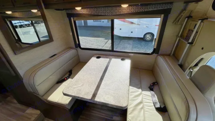 RV rental dinette interior: light-toned benches, table with cup holders, and large windows revealing another RV outside.. Thor Four Winds 2021