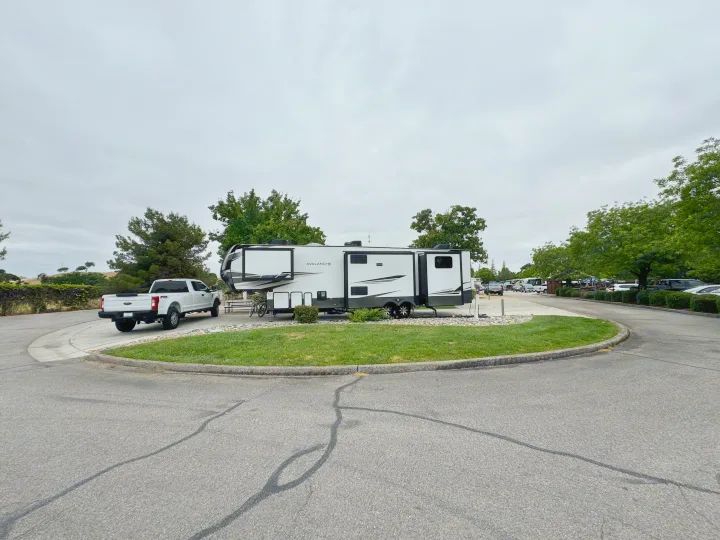 White fifth-wheel RV rental set up at a shaded RV park site with a pickup truck under cloudy skies.. Keystone RV Avalanche 2021