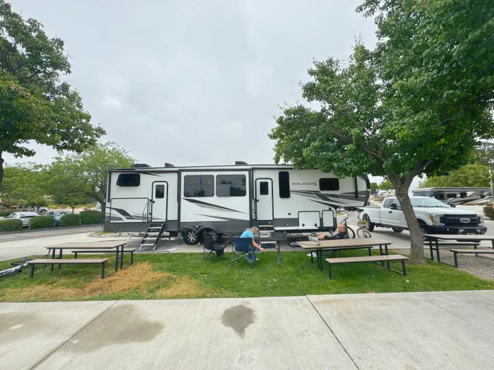 Spacious Avalanche fifth-wheel RV rental parked at a serene campground. Two people relax outdoors near their truck.. Keystone RV Avalanche 2021