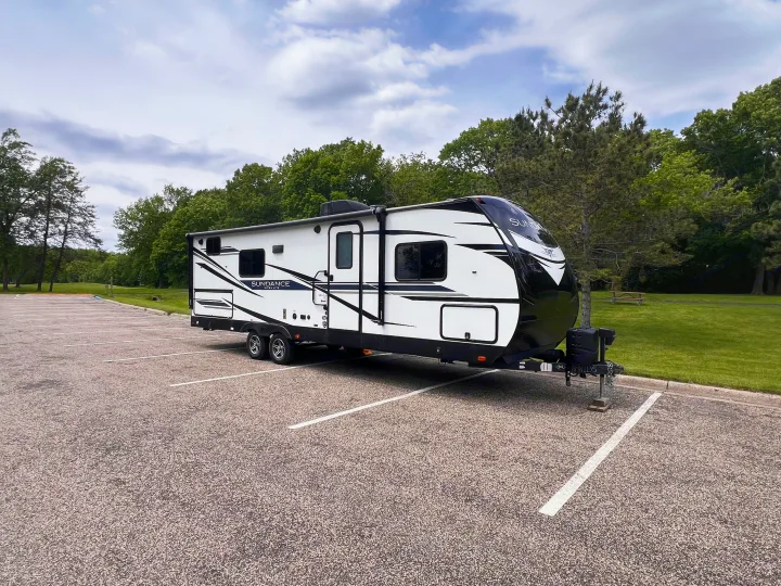 Sleek white and black Sundance Ultra Lite travel trailer RV rental parked in a lot surrounded by trees under a cloudy sky.. Heartland Sundance 2022