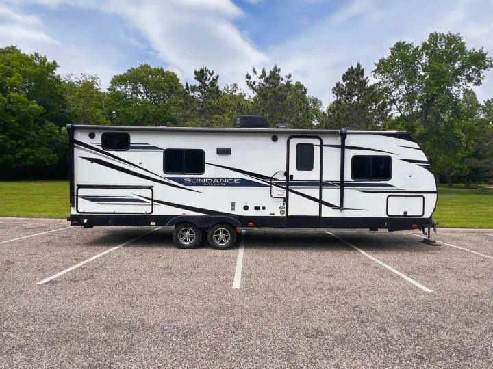 Spacious white travel trailer RV rental, a Sundance Ultra Lite model, parked in a lot with green trees and blue sky.. Heartland Sundance 2022