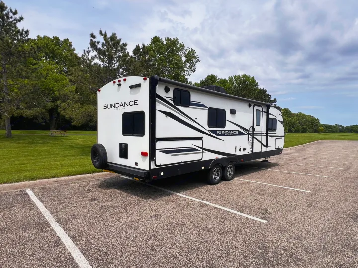 White "Sundance Ultra Lite" travel trailer in a parking lot, green grass and trees behind. Great for an rv rental adventure.. Heartland Sundance 2022