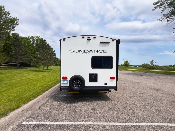 Sundance Ultra Lite travel trailer RV rental, rear view, in parking lot by green trees under cloudy skies.. Heartland Sundance 2022