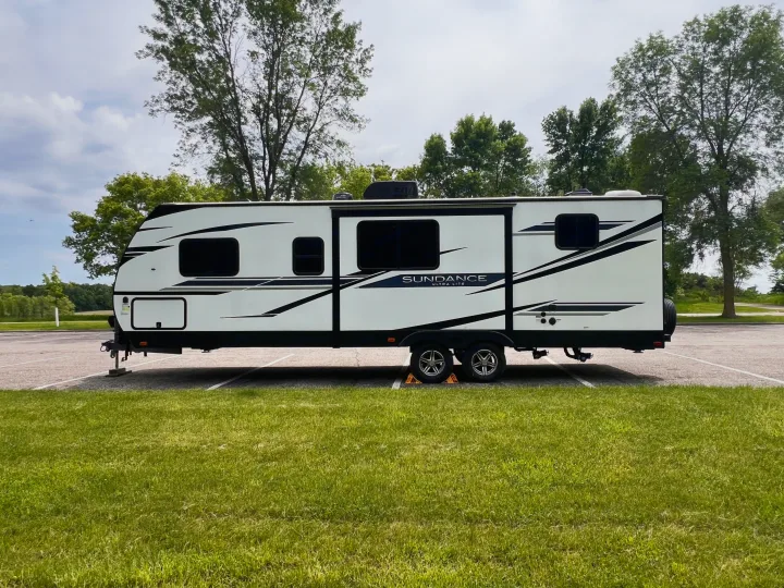 Spotless white Sundance Ultra Lite travel trailer for RV rental, parked on asphalt with grass foreground & trees.. Heartland Sundance 2022