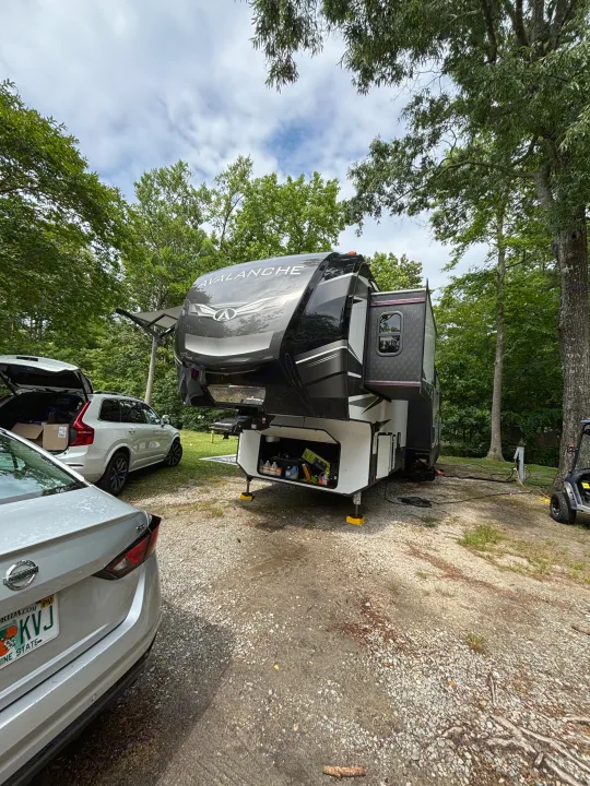 A black and white Avalanche fifth-wheel RV rental, with slide-outs extended, is parked on a gravel campsite amidst green trees. Cars are nearby.. Keystone RV Avalanche 2021