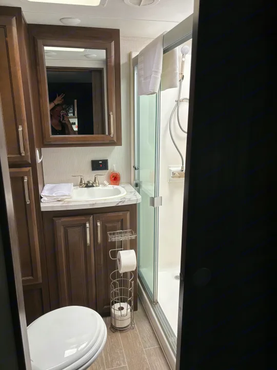 This RV rental bathroom features a dark wood vanity, white sink, toilet, glass shower, and a cheerful selfie in the mirror.. Keystone RV Avalanche 2021