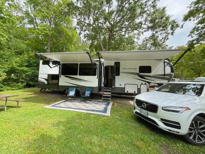 Spacious RV rental set up at a green campsite with a white Volvo, picnic area, and extended awnings for shade.. Keystone RV Avalanche 2021