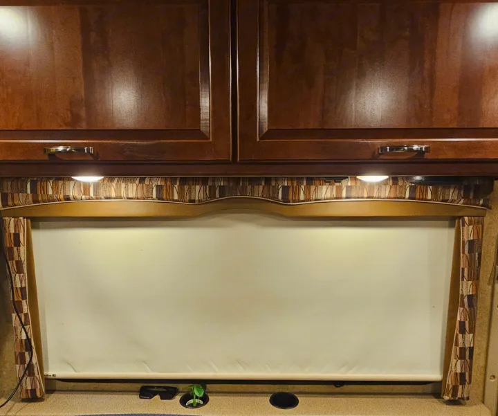 Inside an RV rental: dark wood cabinets with bright under-cabinet lights, a patterned valance, and a lowered window blind. A small plant sits below.. Thor Chateau 2016