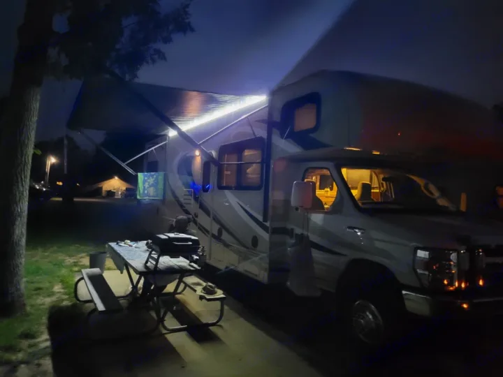 Enjoying the night in an RV rental! Bright awning lights illuminate a cozy campsite with a picnic table & grill.. Thor Chateau 2016