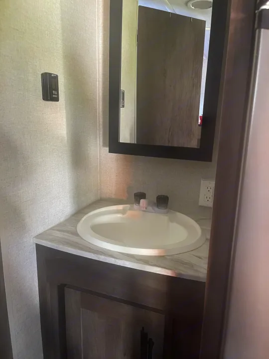 Compact RV rental bathroom featuring a sink, faucet, mirrored cabinet, wall thermostat, and electrical outlet.. Forest River Wildwood X-Lite 2021