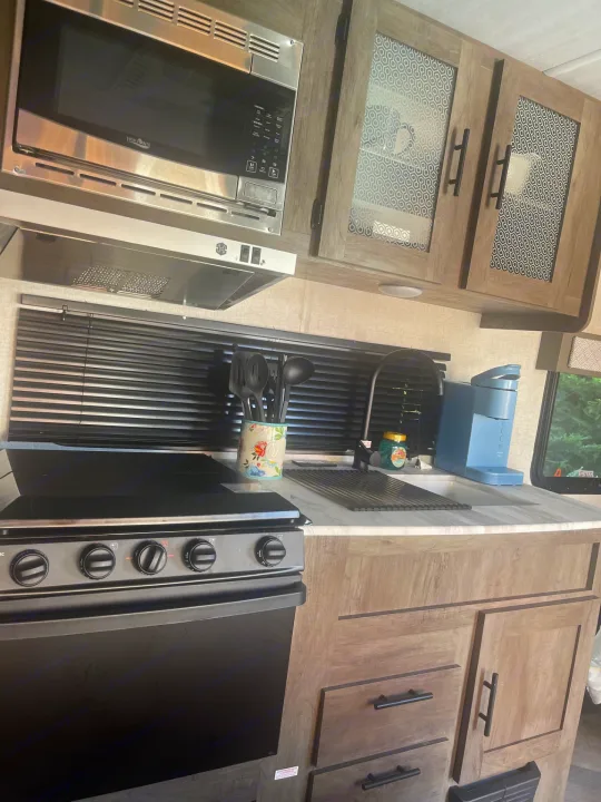 Well-equipped RV rental kitchen: stainless microwave, black stove/oven, sink, and wood-grain cabinets with patterned glass.. Forest River Wildwood X-Lite 2021