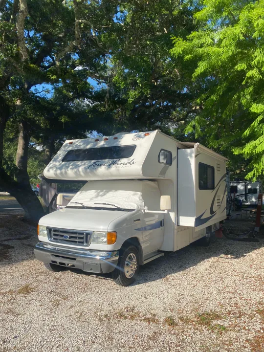 Sunny view of a white Ford Class C Four Winds RV rental parked on gravel at a lush, shaded campground site.. Four Winds 5000 2008