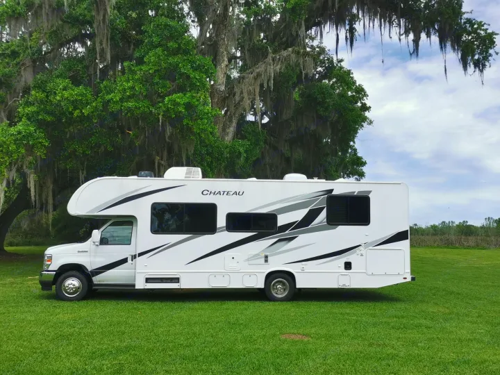 A white Chateau RV rental is parked on a green field with large trees draped in Spanish moss. Ideal for a scenic adventure.. Thor Motor Coach Chateau 2024