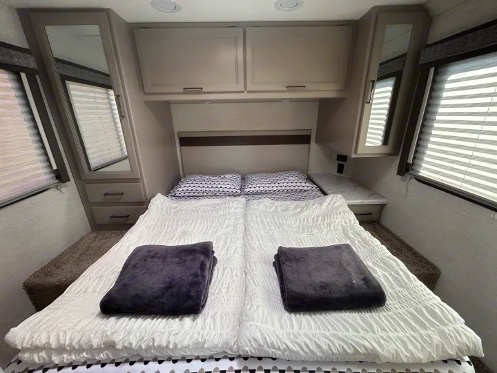 Comfortable RV rental bedroom with a bed, white bedding, two gray blankets, mirrored wardrobes, and overhead cabinets.. Thor Motor Coach Chateau 2024