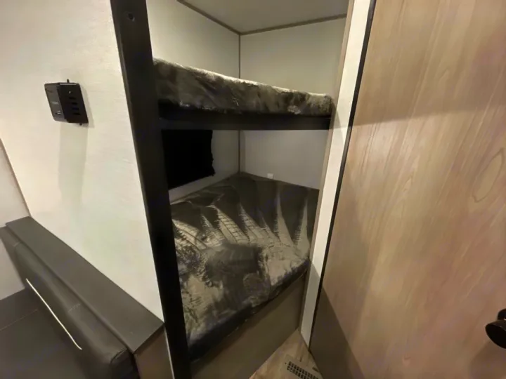 RV rental interior: bunk beds with gray bedding, a gray bench with white stripe, wall USB outlet, and a light wood-grain door.. Coleman LT 274BH 2024