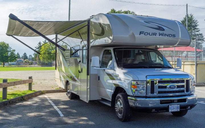 Light-colored Four Winds Class C RV rental parked with awning extended in a park setting on a sunny day.. Thor Four Winds 2019