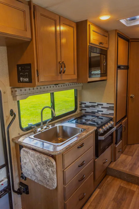 RV rental kitchen interior featuring a sink, stove, microwave, ample cabinetry, and a bright window view.. Thor Four Winds 2019