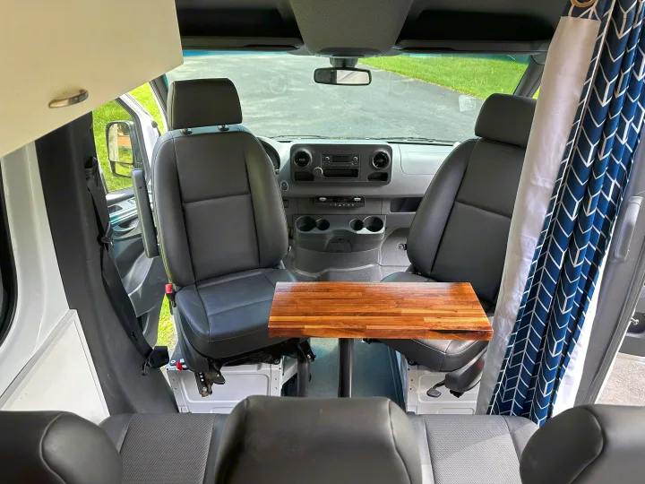 Spacious camper van interior, featuring front seats, a central wooden table, and blue patterned curtains for an RV rental.. Mercedes-Benz Sprinter 2021