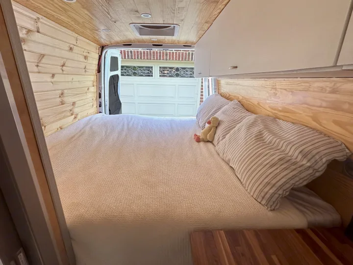 Cozy camper van interior for RV rental. Features a spacious bed, wood paneling, and overhead storage, open to a garage view.. Mercedes-Benz Sprinter 2021