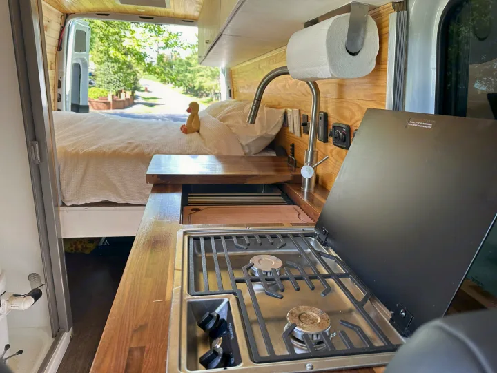 RV rental interior: compact kitchen with stove, sink, and wood counters, leading to a comfy bed with a view outside.. Mercedes-Benz Sprinter 2021