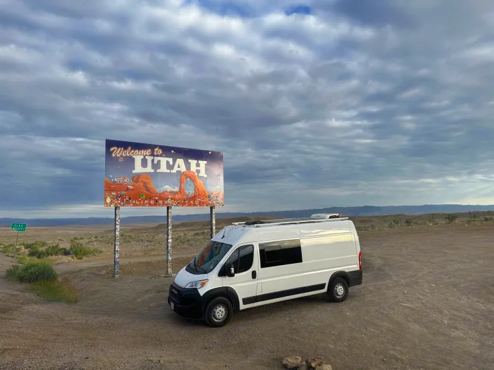 A white camper van, parked on dirt, faces a colorful "Welcome to Utah" sign with an arch graphic. Great for an rv rental trip!. RAM PROMASTER 2500 2023