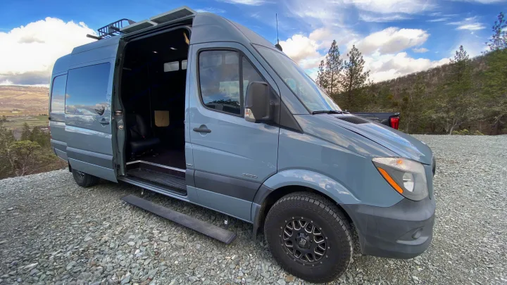 Grey Sprinter camper van on gravel, door open, with scenic hills. This custom van is ready for your RV rental adventure!. Mercedes-Benz Sprinter 2014