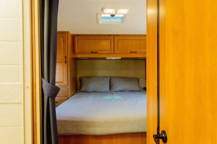 View into a compact RV rental bedroom, featuring a comfy bed with pillows, overhead cabinets, and ceiling lighting.. Thor Majestic 2015