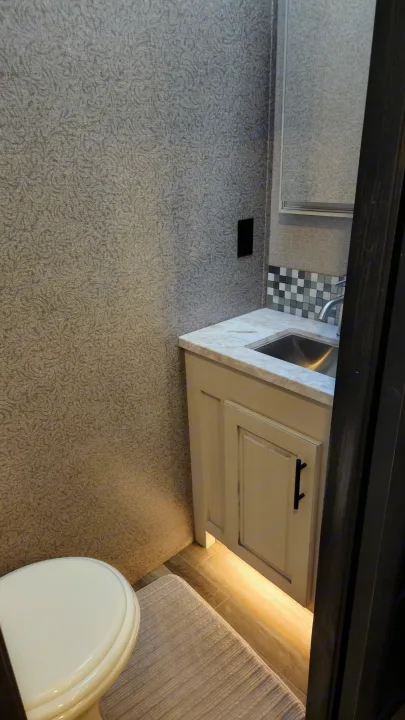 Inside an RV rental bathroom: gray patterned walls, compact vanity with sink, mirror, toilet, and under-cabinet lighting.. Jayco North Point 2021