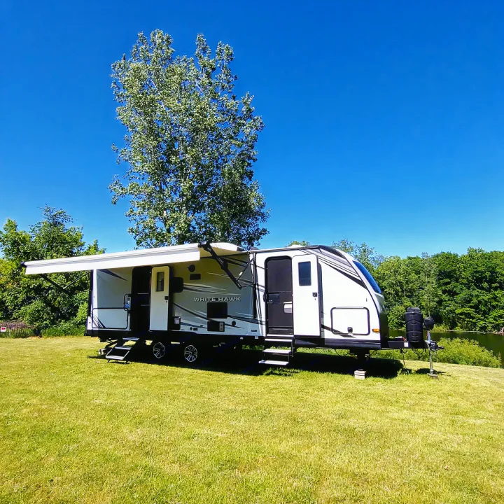 Enjoy a scenic rv rental camping trip! This White Hawk travel trailer is parked on lush grass by a lake, with an awning extended.. Jayco White Hawk 2021