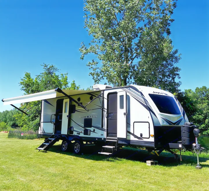White Jayco White Hawk RV rental, awning extended, parked on green grass under a clear blue sky, with lush trees in the background.. Jayco White Hawk 2021