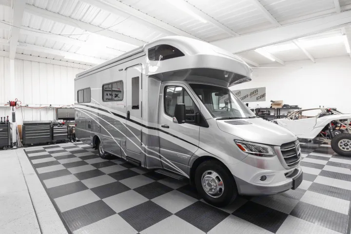 Luxury silver RV rental motorhome parked on checkered flooring in a bright, modern garage with workshop tools and an off-road vehicle.. Winnebago View 2023