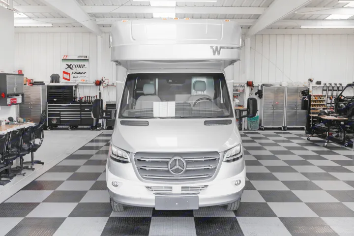 A bright silver Mercedes-Benz Sprinter RV rental, seen from the front, in a clean, organized garage with checkered flooring.. Winnebago View 2023