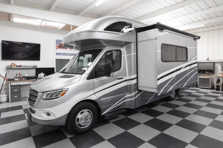 A modern, two-tone gray RV with its large side extended, parked indoors on a black and gray checkered floor. Great for rv rental.. Winnebago View 2023
