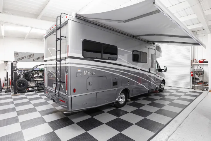 Gray RV rental with awning extended, ready for service in a bright, organized workshop with checkered flooring.. Winnebago View 2023