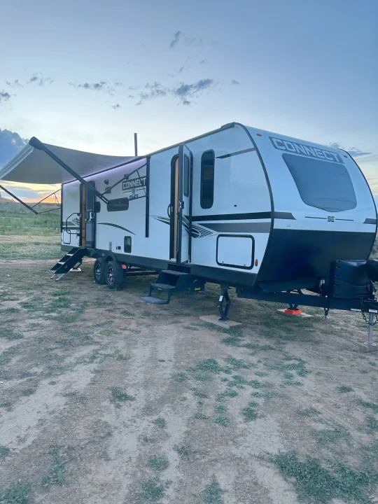 Experience outdoor adventures! This white Connect SE travel trailer, available as an RV rental, is parked with awning extended at dusk.. Connect SE 2022