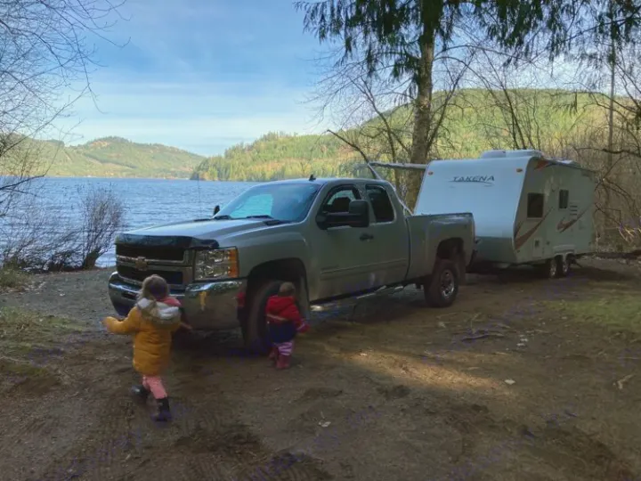 Enjoying nature: A gray pickup and white RV rental by a lake, with two small children playing in the foreground.. Takena Takena Trailer 2010